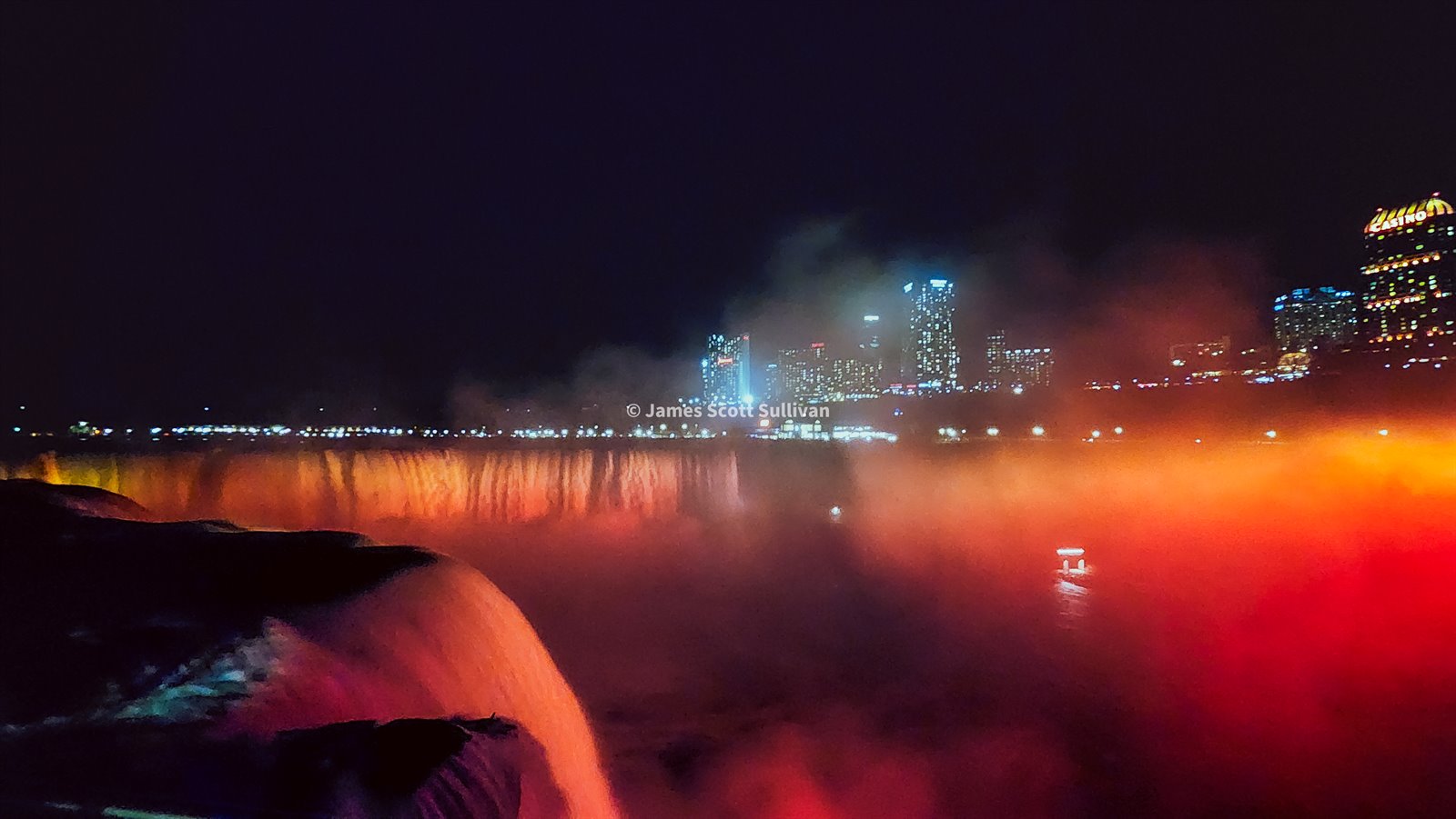 Niagara Falls glowing at night with city lights in the background.